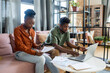 © pressmaster - Young black woman sitting on sofa and looking through financial paper while African man looking at laptop display