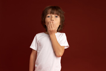 Little boy learned secret and trying to keep it covering mouth with hand, having surprised and shocked face expression, isolated on red studio wall with copy space for your advertisement