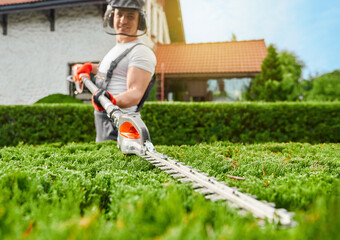 Competent caucasian gardener wearing uniform, safety mask and gloves cutting overgrown bushes with electric trimmer. Concept of people and landscaping.