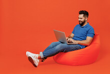 Full Body Young Smiling Happy Caucasian Man Wear Basic Blue T-shirt Sit In Bag Chair Hold Use Work On Laptop Pc Computer Isolated On Plain Orange Background Studio Portrait. People Lifestyle Concept.