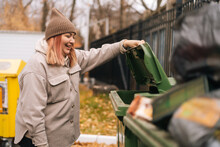 Side View Of Laughing Overweight Female Wearing Autumn Hat And Jacket Opening Trash Can And Looking Inside. Attractive Young Homeless Female Looking For Food In Street Garbage Can In Cloudy Day.