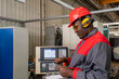 © RGtimeline - CNC Machine Operator In Protective Workwear Checking Measurements With Vernier Caliper. Portrait Of African American Worker In Red Helmet, Hearing Protectors And Work Uniform Next To CNC Controller.