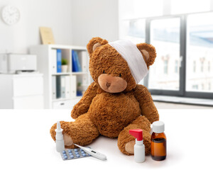 Wall Mural - medicine, healthcare and childhood concept - teddy bear toy with bandaged head thermometer and drugs over medical office at hospital background
