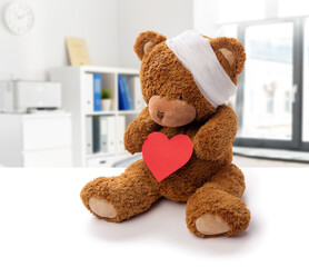 Wall Mural - medicine, healthcare and childhood concept - teddy bear toy with bandaged head holding red heart over hospital background