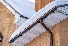 Corner Of House With Roof Made Of Brown Metal Tiles And Gutter Covered With Thick Layer Of Snow In Winter. Metal Downpipe System, Guttering System, External Downpipes And Drainage Pipes