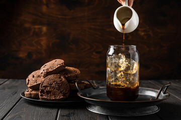 Wall Mural - Making iced coffee. Pouring black coffee from a milk jug into a glass. Chocolate cookies on a plate.