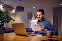 Fatherhood And Remote Business. A Happy Young Dad Works From Home Holds His Sleepy Baby Girl In His Hands, Goes Through Paperwork And Has An Online Meeting On His Laptop With His Colleagues.