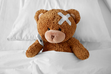 Wall Mural - medicine, healthcare and childhood concept - ill teddy bear toy with medical patch on head and thermometer lying in bed