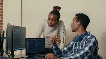 Database developer removing glasses while explaining algorithm to colleague coder pointing at running code on multiple computer screens. System engineers collaborating on development group project.