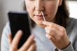 © Syda Productions - medicine, quarantine and pandemic concept - close up of woman with swab and smartphone taking sample from her nose and making nasal coronavirus self test at home
