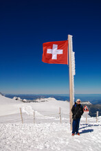 A Caucasion Female Wearing Sun Glasses Standing Under The Swiss Flag At Jungfrau, At The Top Of Europe, On Snow And With Deep Blue Sky Behind