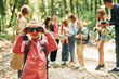 © standret - Girl looking into binoculars. Kids in green forest at summer daytime together