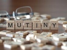 Mutiny Concept Represented By Wooden Letter Tiles On A Wooden Table With Glasses And A Book