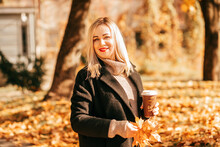 Coffee Break In Sunny Autumn Park. Young Woman Entrepreneur Holds Autumn Leaves And Cup Of Cappuccino Coffee In Her Hands . Walking Alone In Fresh Air. Happy People. Life In Moment.