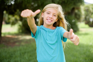 Wall Mural - Caucasian little kid girl wearing blue T-shirt standing outdoors approving doing positive gesture with hand, thumbs up smiling and happy for success. Winner gesture.