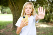 © Roquillo - Caucasian little kid girl wearing white T-shirt standing outdoors using and texting with smartphone with open hand doing stop sign with serious and confident expression, defense gesture