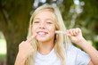 © Roquillo - Caucasian little kid girl wearing whiteT-shirt standing outdoors holding an invisible aligner and pointing to her perfect straight teeth. Dental healthcare and confidence concept.