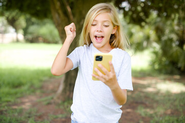 Wall Mural - Portrait of Caucasian little kid girl wearing white T-shirt standing outdoors holding in hands cell celebrating