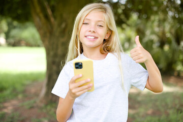 Wall Mural - Portrait of Caucasian little kid girl wearing white T-shirt standing outdoors using and texting with smartphone  happy with big smile doing ok sign, thumb up with fingers, excellent sign