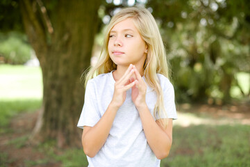 Wall Mural - Caucasian little kid girl wearing whiteT-shirt standing outdoors steepls fingers and looks mysterious aside has great evil plan in mind