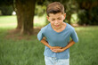 Caucasian little kid boy wearing blue T-shirt standing outdoor with hand on stomach because nausea, painful disease feeling unwell. Ache concept.