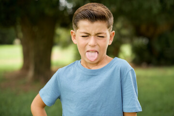 Wall Mural - Caucasian little kid boy wearing blue T-shirt standing outdoor sticking tongue out happy with funny expression. Emotion concept.