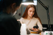 © dikushin - Close-up view from back to happy young woman typing online message on mobile phone during professional manicure master removing gel polish from nails, using manicure machine.