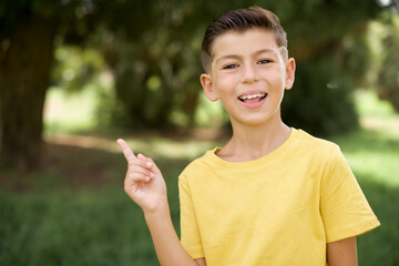 Wall Mural - Caucasian little kid boy wearing yellow T-shirt standing outdoors laughs happily points away on blank space demonstrates shopping discount offer, excited by good news or unexpected sale.