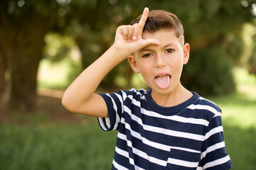 Funny beautiful Caucasian little kid boy wearing stripped T-shirt standing outdoors makes loser gesture mocking at someone sticks out tongue making grimace face.