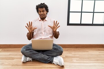 Canvas Print - African man with curly hair using laptop sitting on the floor afraid and terrified with fear expression stop gesture with hands, shouting in shock. panic concept.