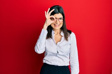 Wall Mural - Young hispanic woman wearing business shirt and glasses doing ok gesture with hand smiling, eye looking through fingers with happy face.