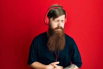 Poster - Redhead man with long beard listening to music using headphones with hand on stomach because indigestion, painful illness feeling unwell. ache concept.