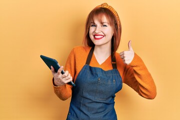 Sticker - Redhead young woman wearing waitress apron holding touchpad device taking order smiling happy and positive, thumb up doing excellent and approval sign