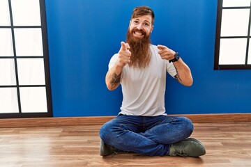 Sticker - Redhead man with long beard sitting on the floor at empty room pointing fingers to camera with happy and funny face. good energy and vibes.