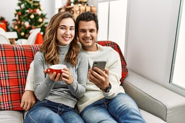 Wall Mural - Young hispanic couple drinking coffee using smartphone sitting on the sofa at home.