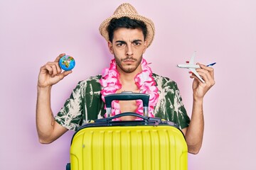 Wall Mural - Young hispanic man wearing summer style and hawaiian lei holding world ball and plane toy skeptic and nervous, frowning upset because of problem. negative person.