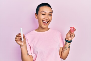 Sticker - Beautiful hispanic woman with short hair holding menstrual cup and tampon winking looking at the camera with sexy expression, cheerful and happy face.