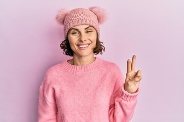 Wall Mural - Young brunette woman wearing cute wool cap showing and pointing up with fingers number two while smiling confident and happy.