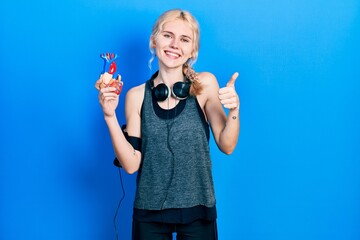 Poster - Beautiful caucasian sports woman with blond hair holding heart smiling happy and positive, thumb up doing excellent and approval sign