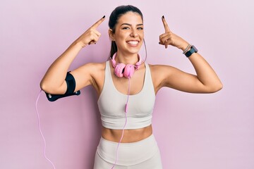 Wall Mural - Young hispanic woman wearing gym clothes and using headphones smiling pointing to head with both hands finger, great idea or thought, good memory