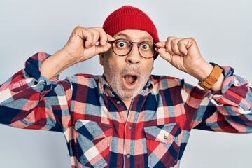 Wall Mural - Handsome mature man holding glasses, wearing wool hat afraid and shocked with surprise and amazed expression, fear and excited face.