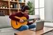 © Krakenimages.com - Young hispanic man having online classical guitar lesson sitting on the sofa at home.