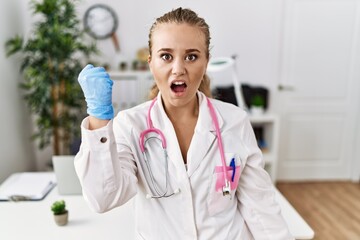 Poster - Young caucasian woman wearing doctor uniform and stethoscope at the clinic angry and mad raising fist frustrated and furious while shouting with anger. rage and aggressive concept.