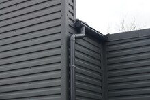 Black Metal Wall Of A Building With A Plastic Drain Pipe On The Street Against A Gray Sky