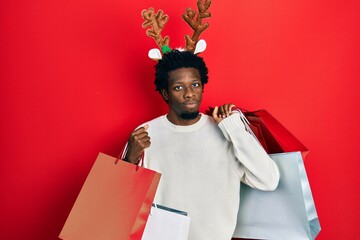 Canvas Print - Young african american man wearing deer christmas hat holding shopping bags skeptic and nervous, frowning upset because of problem. negative person.