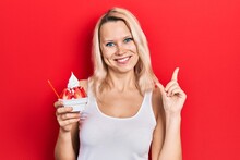 Beautiful Caucasian Blonde Woman Eating Strawberry Ice Cream Smiling Happy Pointing With Hand And Finger To The Side