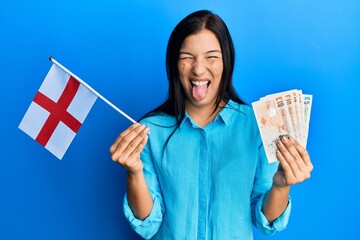 Poster - Young latin woman holding england flag and pounds banknotes sticking tongue out happy with funny expression.