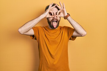 Wall Mural - Caucasian man with beard wearing casual yellow t shirt doing ok gesture like binoculars sticking tongue out, eyes looking through fingers. crazy expression.