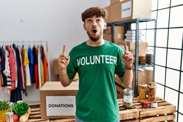 Poster - Young arab man wearing volunteer t shirt at donations stand amazed and surprised looking up and pointing with fingers and raised arms.