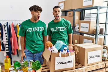 Sticker - Young gay couple wearing volunteer t shirt at donations stand angry and mad screaming frustrated and furious, shouting with anger. rage and aggressive concept.